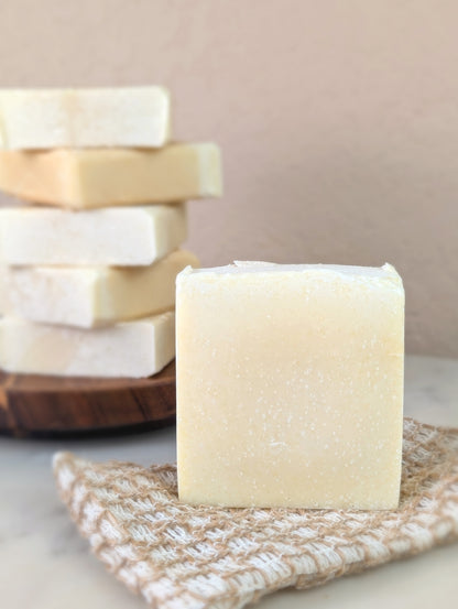 A single while soap bar in the foreground sitting on top of a flat textured soap saver bag with a stack of soap bars on a wooden dish in the background.