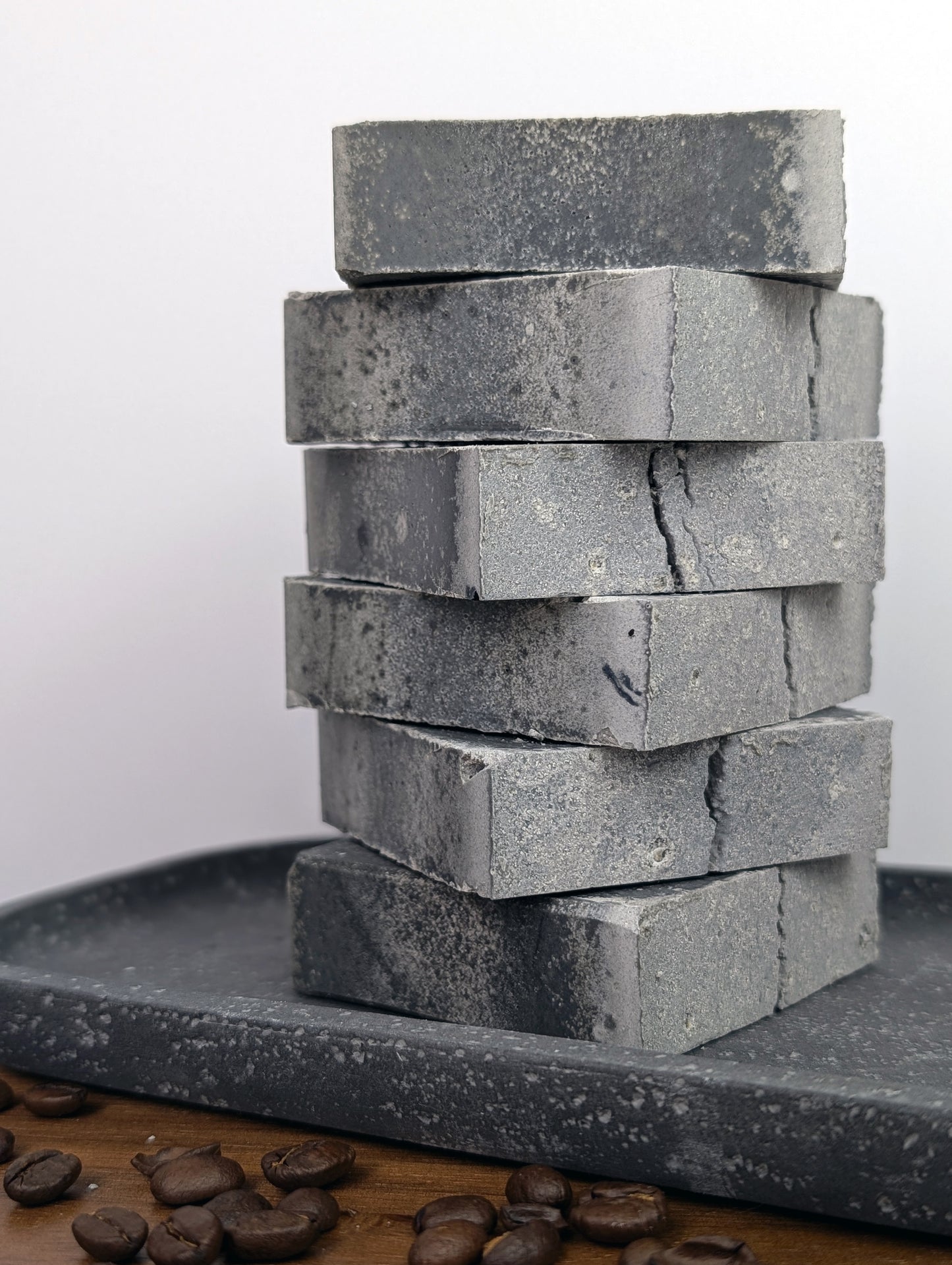 Stacked dark gray soaps on a slate gray dish with coffee beans in the foreground with a white background.