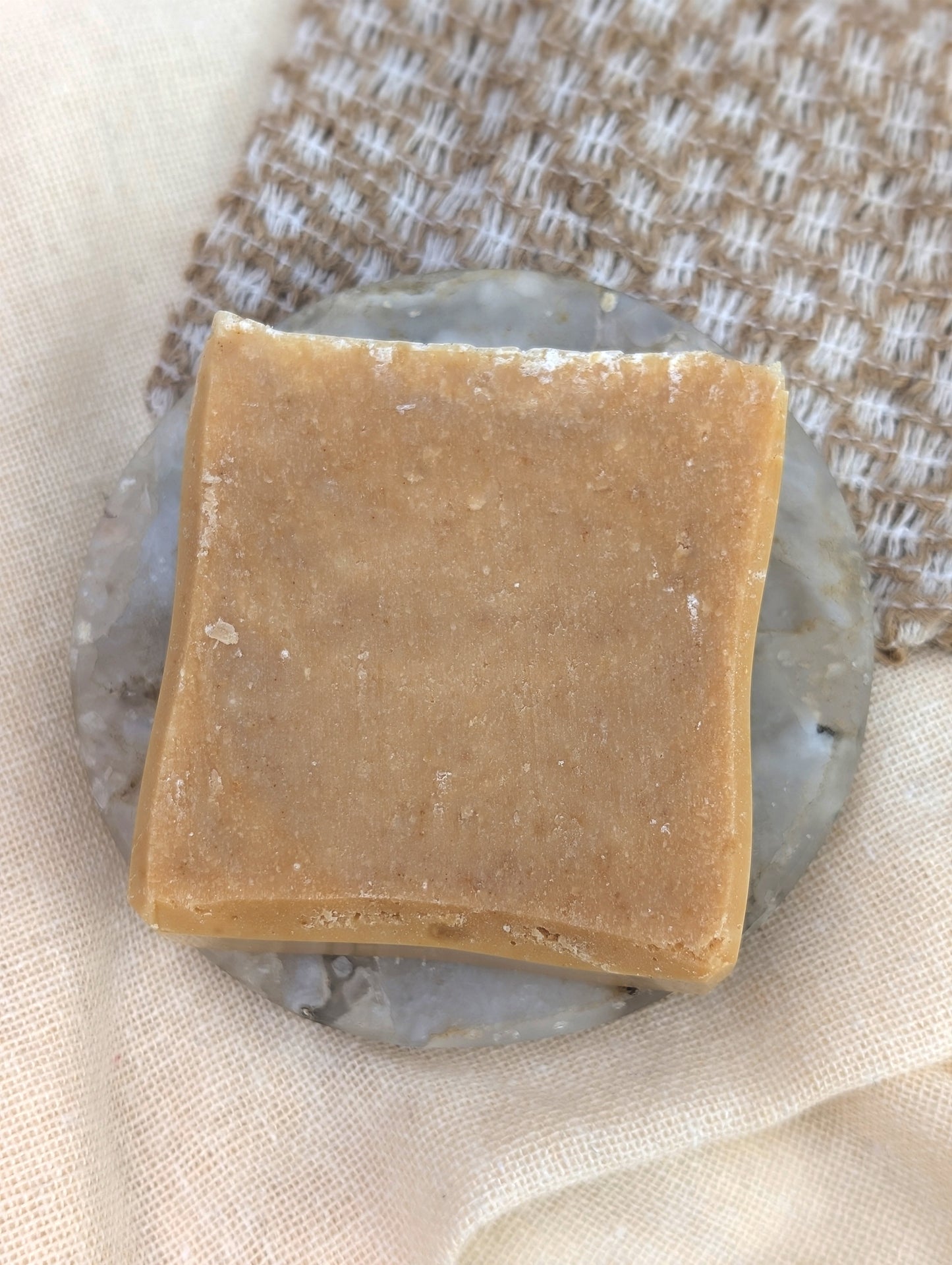 Single beige square soap laid on a round marble dish on top of a textured background.