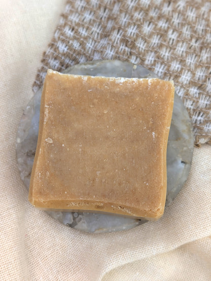 Single beige square soap laid on a round marble dish on top of a textured background.